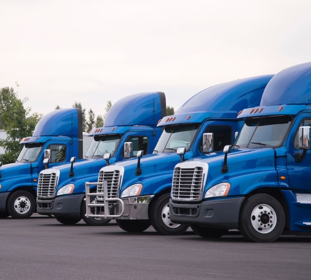 Four blue semi-trucks parked side by side on a paved lot under a cloudy sky.