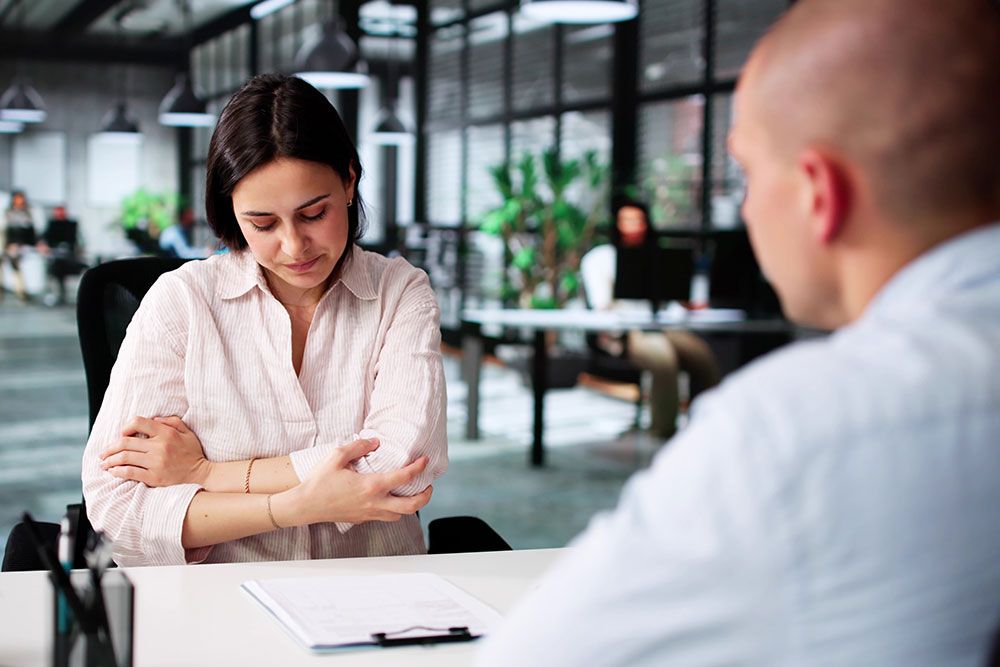 Woman in office setting appears stressed, holding arm during a conversation with a colleague.