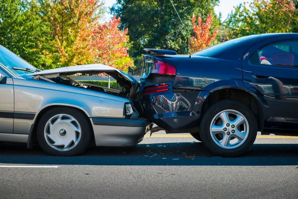 Two cars in a rear-end collision on a sunny road with trees in the background.
