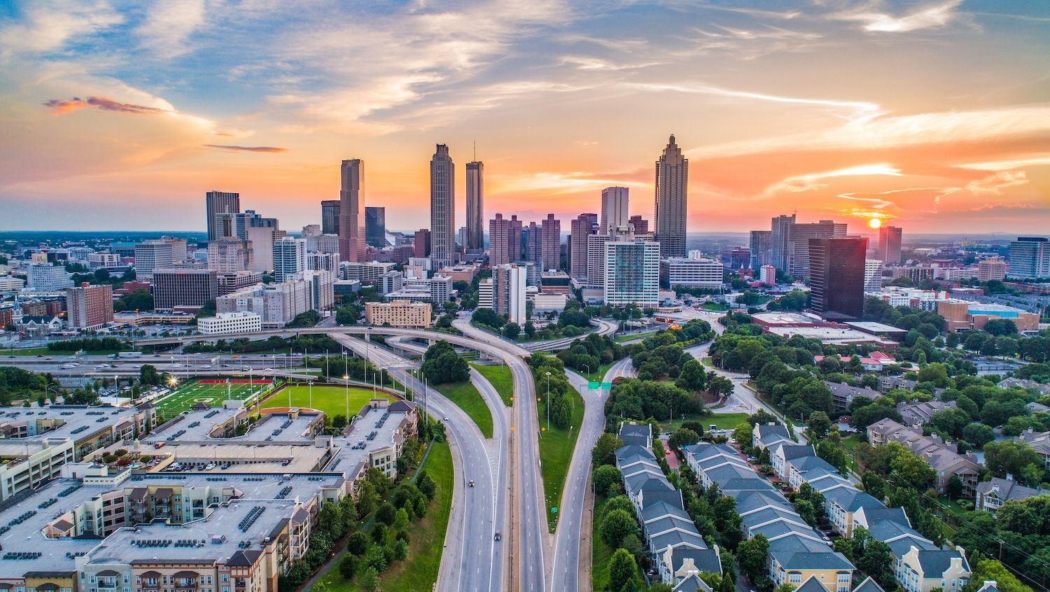 Aerial view of city skyline at sunset, featuring skyscrapers, highways, and residential areas with colorful sky.