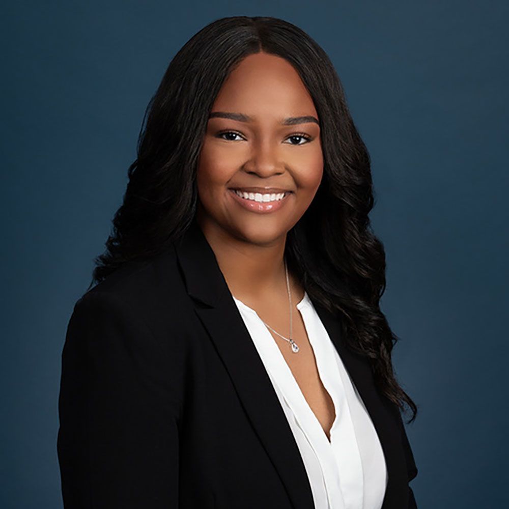 Toni Grier in a black blazer smiling against a blue background. Business portrait for corporate profile.