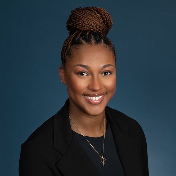Portrait of a smiling person with braided hair against a blue background, wearing a black blazer and necklace.