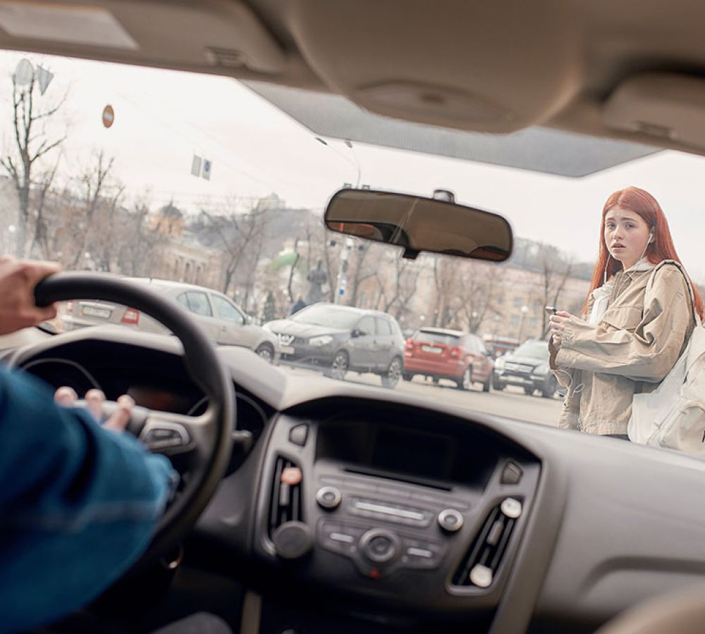 A driver spots a pedestrian crossing a busy street, holding a smartphone and wearing a beige jacket with a white backpack.