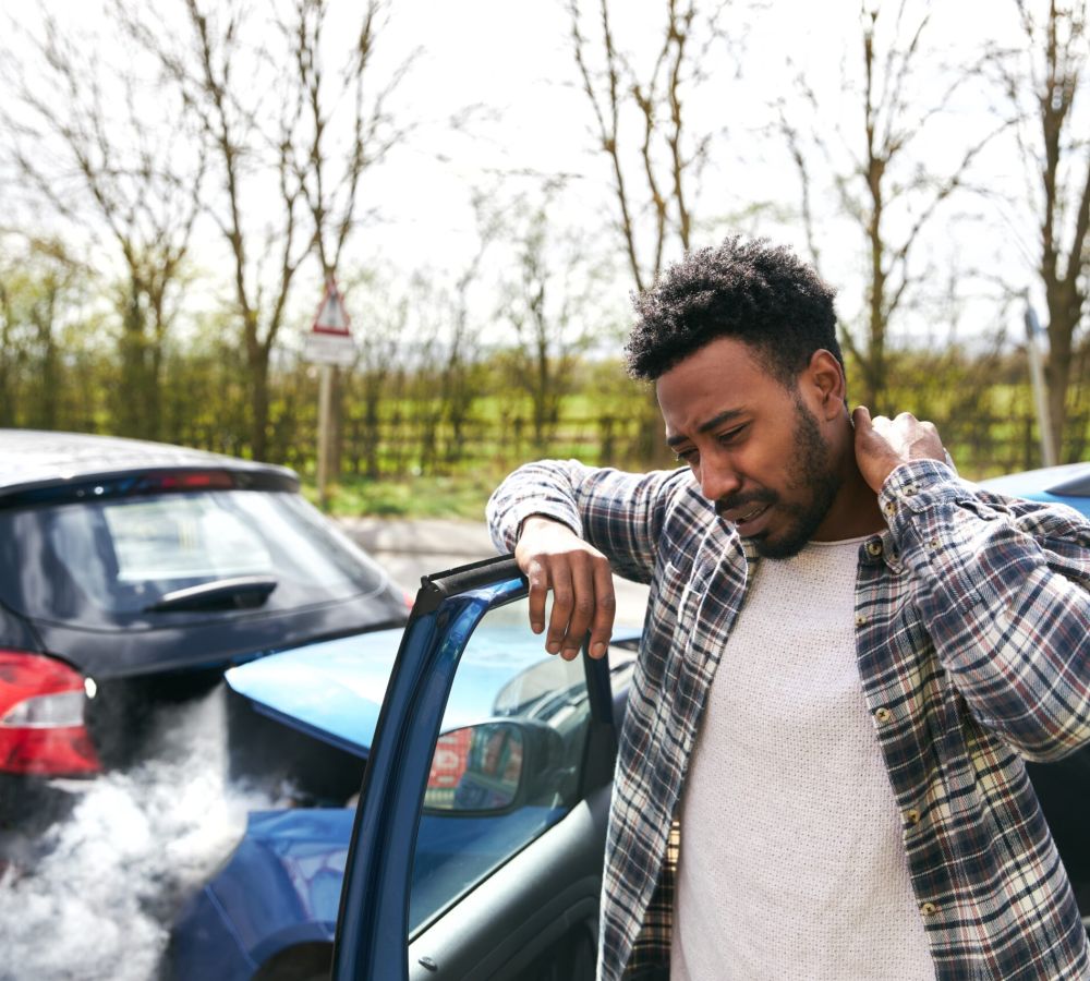 Man holding neck after car accident, examining vehicle damage on road.