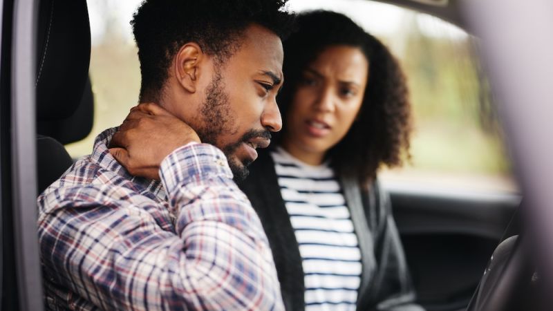 Man in car holding neck, looking pained, while woman beside him looks concerned.